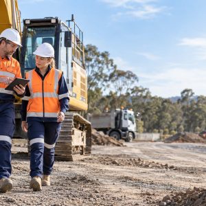 Two workers in hi-vis reviewing a plant risk assessment on a tablet beside a tracked excavator on a civil site